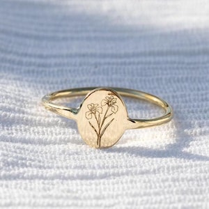 Puede incluir: Un anillo dorado con una cara ovalada con un diseño floral. El anillo tiene una banda delgada y una textura martillada. El diseño floral está grabado en la cara ovalada.