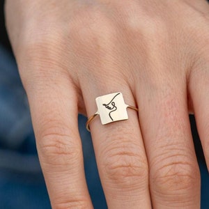 Op de afbeelding: Een gouden ring met een vierkante band met een minimalistische lijntekening van een gezicht.