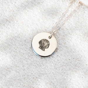 Op de afbeelding: Een zilveren ketting met een ronde hanger met een gedetailleerd schelpontwerp.