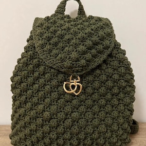 Puede incluir: Una mochila de ganchillo color verde oliva con un patrón texturizado de puntos de burbuja en todo el cuerpo y la solapa. La bolsa incluye un asa superior y un cierre metálico dorado en forma de corazón centrado en la solapa frontal.