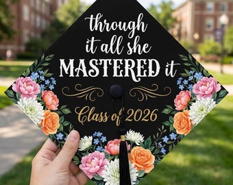 Peut inclure: Coiffe de graduation noire avec un motif floral et le texte "through it all she MASTERED it Class of 2026". La coiffe présente des fleurs roses, oranges, blanches et bleues. Une gland noir pend du centre.