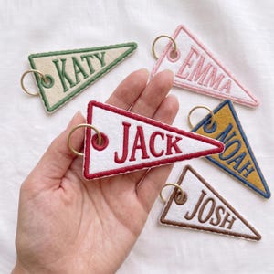 May include: Five triangular felt pennant keychains with gold-tone metal rings. Each features embroidered names: KATY in green, EMMA in pink, JACK in red, NOAH in blue on a mustard background, and JOSH in brown on a white background with brown trim.