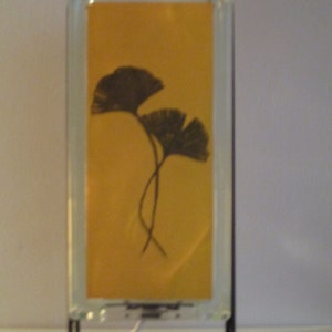 K&ouml;nnte beinhalten: Rechteckige Glaslampe mit gelbem Hintergrund und schwarzem Ginkgo-Blatt-Design. Die Lampe hat einen schwarzen Metallst&auml;nder und ein wei&szlig;es Netzkabel. Das Design ist schlicht und elegant.