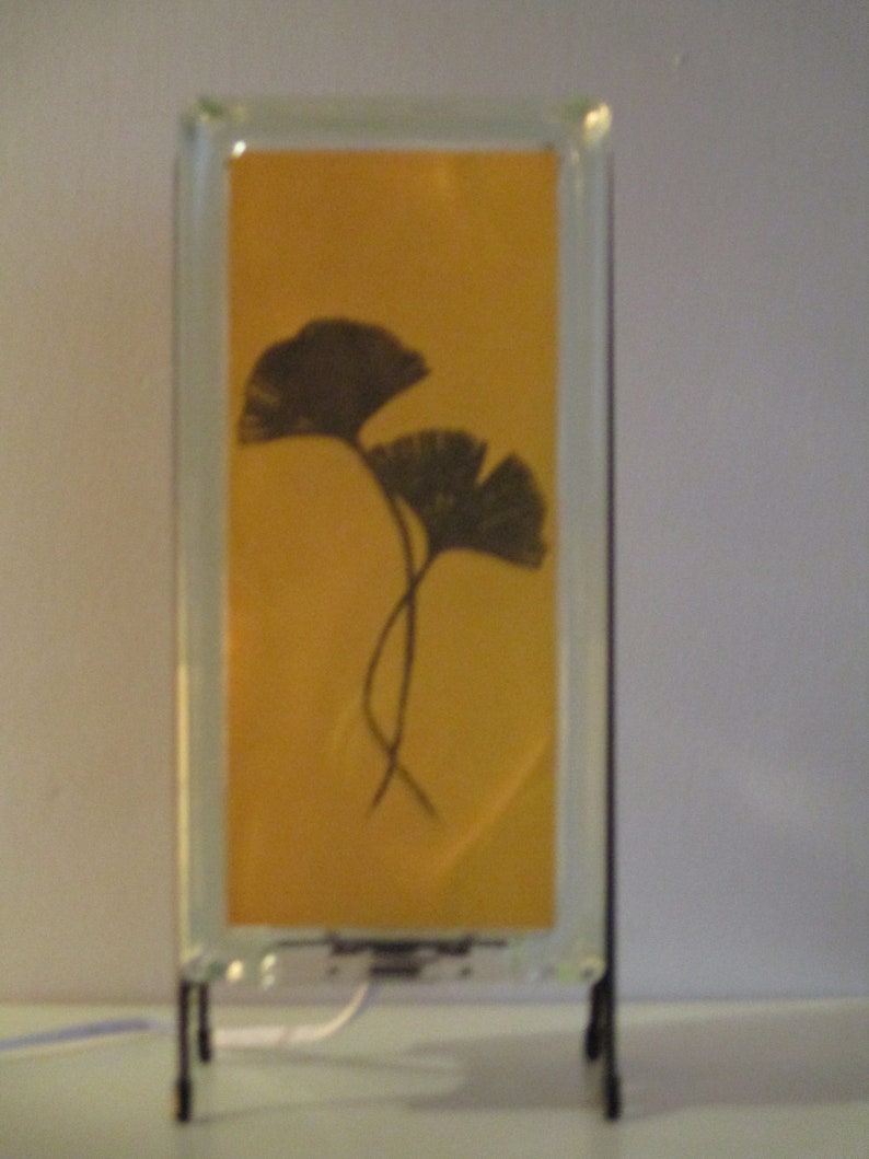 K&ouml;nnte beinhalten: Rechteckige Glaslampe mit gelbem Hintergrund und schwarzem Ginkgo-Blatt-Design. Die Lampe hat einen schwarzen Metallst&auml;nder und ein wei&szlig;es Netzkabel. Das Design ist schlicht und elegant.