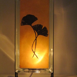 K&ouml;nnte beinhalten: Eine rechteckige Tischlampe mit einem transparenten Glasrahmen und einem warmen, bernsteinfarbenen Innenraum. Der Innenraum zeigt eine schwarze Silhouette von zwei Ginkgobl&auml;ttern. Die Lampe wird von einem schwarzen Metallst&auml;nder getragen und strahlt ein weiches, warmes Licht aus.