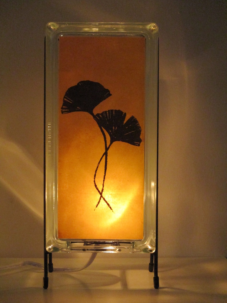 K&ouml;nnte beinhalten: Eine rechteckige Tischlampe mit einem transparenten Glasrahmen und einem warmen, bernsteinfarbenen Innenraum. Der Innenraum zeigt eine schwarze Silhouette von zwei Ginkgobl&auml;ttern. Die Lampe wird von einem schwarzen Metallst&auml;nder getragen und strahlt ein weiches, warmes Licht aus.