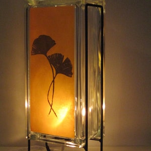 K&ouml;nnte beinhalten: Dekorative Lampe mit einem transparenten Glasblock-Design und einem warmen orangefarbenen Hintergrund. Der Hintergrund zeigt eine Silhouette von zwei Ginkgobl&auml;ttern. Die Lampe wird von einem schwarzen Metallrahmen getragen und strahlt ein weiches, einladendes Licht aus.