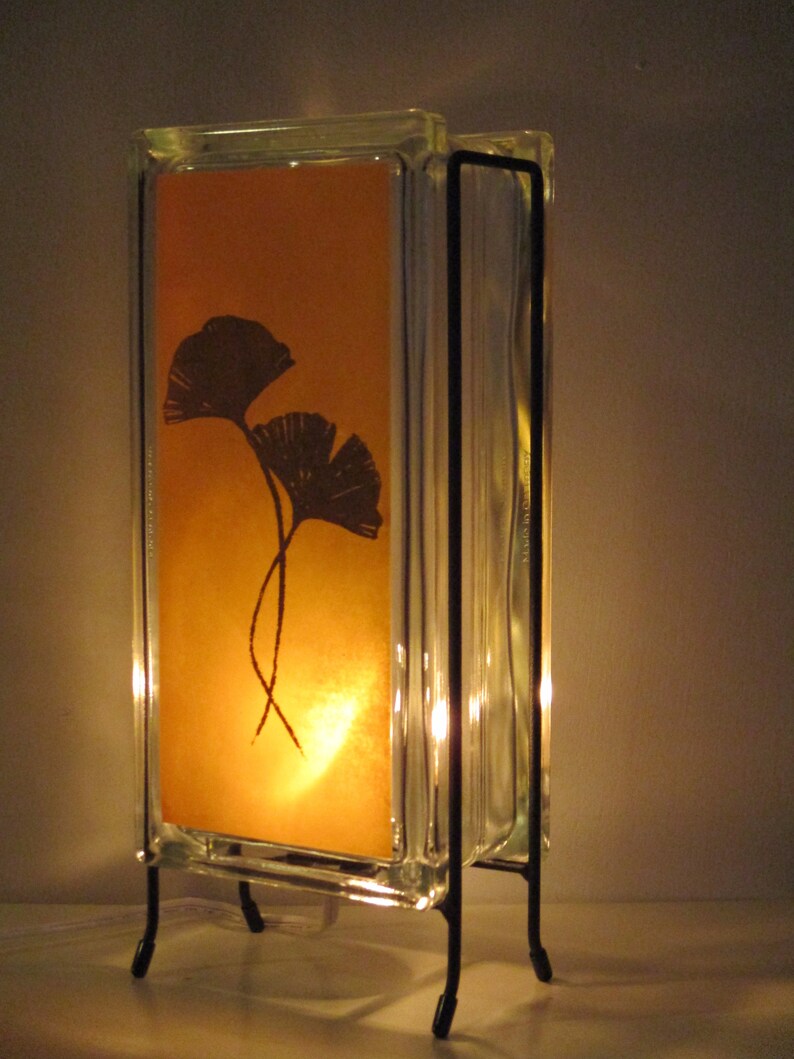 K&ouml;nnte beinhalten: Dekorative Lampe mit einem transparenten Glasblock-Design und einem warmen orangefarbenen Hintergrund. Der Hintergrund zeigt eine Silhouette von zwei Ginkgobl&auml;ttern. Die Lampe wird von einem schwarzen Metallrahmen getragen und strahlt ein weiches, einladendes Licht aus.