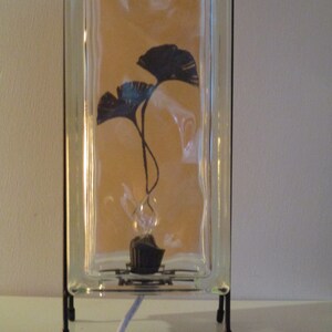 K&ouml;nnte beinhalten: Dekorative Glasblocklampe mit schwarzem Metallrahmen. Im Inneren des Glasblocks befindet sich eine Silhouette von zwei dunkelblauen Ginkgobl&auml;ttern. Die Lampe hat einen schwarzen Sockel und ein wei&szlig;es Netzkabel.