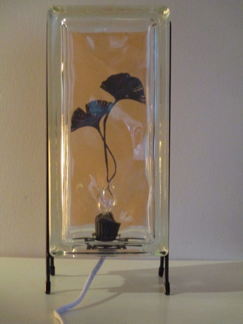 K&ouml;nnte beinhalten: Dekorative Glasblocklampe mit schwarzem Metallrahmen. Im Inneren des Glasblocks befindet sich eine Silhouette von zwei dunkelblauen Ginkgobl&auml;ttern. Die Lampe hat einen schwarzen Sockel und ein wei&szlig;es Netzkabel.