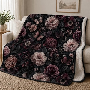 May include: A floral throw blanket featuring a dark background with large, detailed flowers in shades of pink, purple, and cream. Butterflies are scattered throughout the design. The blanket has a soft, white sherpa lining.