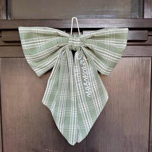 May include: A sage green and white plaid fabric bow featuring a central knot and two long, pointed tails. The right tail is embroidered with the white text "The Swathson Family" in a cursive script. A white cord loop is attached to the top for hanging.