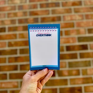 May include: A rectangular notepad with a blue border and a white center. The top features a pink spiral binding graphic. The text reads things i need to OVERTHINK in blue and pink, with the word SWAM CO. printed in small blue letters at the bottom.