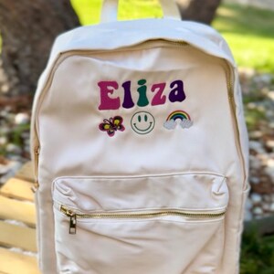 May include: Cream-colored backpack with gold zippers and a front pocket. The backpack features the name "Eliza" embroidered in colorful letters, along with a butterfly, smiley face, and rainbow design. The bag has a top handle and adjustable straps.