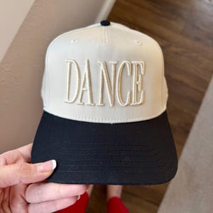 May include: A baseball cap with a cream-colored crown and a black brim. The word "DANCE" is embroidered in a raised, beige font across the front of the cap. The cap has a curved brim and a small, black button on top.