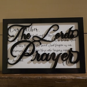 May include: A framed wall art piece featuring the Lord's Prayer. The black frame surrounds a white background with the prayer's text. The words "The Lord's Prayer" are cut out in a stylish, cursive font, adding a decorative element.