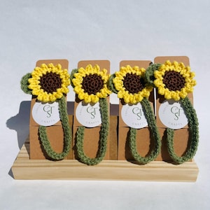 May include: Four handmade crochet sunflower bookmarks. Each bookmark features a yellow sunflower with a brown centre and a green stem. The bookmarks are displayed on a wooden stand with a brown background. Each bookmark has a white label with the text "Crafts".