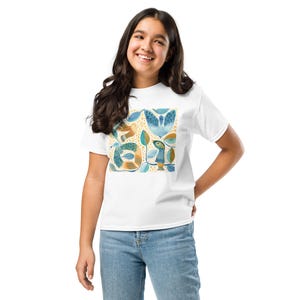 Könnte beinhalten: Ein weißes kurzärmeliges T-Shirt mit einem zentralen Grafikdruck aus stilisierten Vögeln und Blättern in Blau-, Petrol- und Senfgelbtönen vor einem Hintergrund aus kleinen gelben Punkten, kombiniert mit einer blauen Jeanshose.