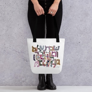 K&ouml;nnte beinhalten: Eine wei&szlig;e Tragetasche mit schwarzen Schulterriemen und einem farbenfrohen hebr&auml;ischen Grafikdruck. Das Design besteht aus stilisierten hebr&auml;ischen Buchstaben, die mit verschiedenen komplizierten Mustern in Rosa-, Gr&uuml;n-, Braun- und Blaut&ouml;nen gef&uuml;llt sind.