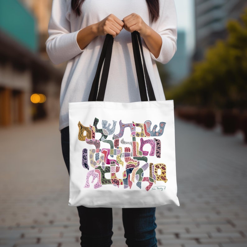 K&ouml;nnte beinhalten: Eine wei&szlig;e Tragetasche mit schwarzen Henkeln und einem bunten, abstrakten hebr&auml;ischen Kalligrafie-Design. Das Kunstwerk besteht aus miteinander verbundenen, gemusterten Formen in Rosa, Lila, Blau, Gelb und Gr&uuml;n, die hebr&auml;ischen Text bilden.