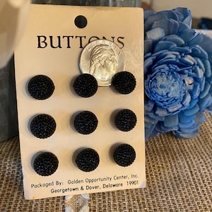 Puede incluir: Una tarjeta con la palabra "BUTTONS" impresa en ella. Hay ocho botones negros con una superficie texturizada dispuestos en dos filas de cuatro. La tarjeta también incluye el texto "Packaged By: Golden Opportunity Center, Inc. Georgetown & Dover, Delaware 19901".