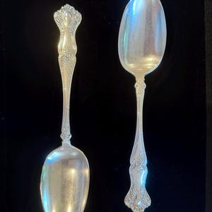 May include: Two silver-toned metal spoons featuring an ornate, embossed floral and vine pattern along the handles. The spoons have polished, oval-shaped bowls and detailed, decorative stems that taper toward the base of the handle.
