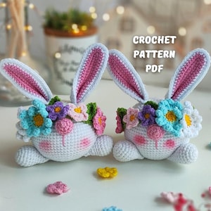 以下が含まれることがあります： ピンクの裏地が付いた耳と花の冠が付いた、2つの白いかぎ針編みのウサギのフィギュア。 ウサギは笑顔で、カラフルな花で飾られています。 テキスト「CROCHET PATTERN PDF」が表示されます。