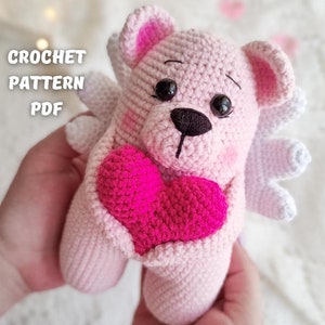 Puede incluir: Un oso de peluche rosa tejido a mano con un corazón rosa brillante. El oso tiene alas blancas y ojos negros. El texto "CROCHET PATTERN PDF" es visible. Un artículo hecho a mano encantador.