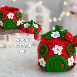 Puede incluir: Patrón de crochet PDF para un contenedor con forma de fresa con una puerta verde y flores blancas. El contenedor está hecho de hilo rojo y tiene una tapa verde con una fresa en la parte superior.