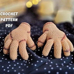 以下が含まれることがあります： 縫い付けられた茶色の編みぐるみ手袋のペアを作るための、かぎ針編みパターンのPDF。手袋は黒と白のドット柄の布の上に置かれています。