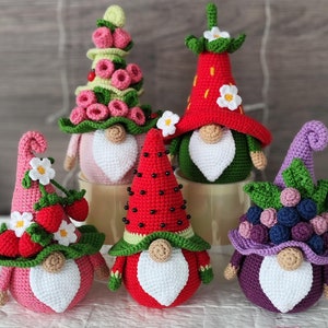 May include: Five crocheted gnomes with different fruit-themed hats. The gnomes are pink, red, and purple. The hats are decorated with flowers and fruit.