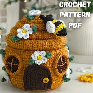 Könnte beinhalten: Gehäkelte Bienenstock-förmige Box in Senfgelb mit Deckel, verziert mit einer Hummel und weißen Blumenakzenten. Die Box hat eine dunkelbraune Tür und runde Fenster. Der Text "CROCHET PATTERN PDF" ist sichtbar.