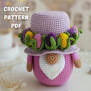 May include: Crochet pattern PDF for a purple and pink gnome with a flower crown. The gnome is wearing a purple hat with yellow and purple flowers and green leaves.