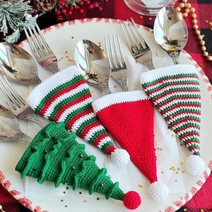 May include: A white plate with four crocheted Christmas-themed cutlery holders. The holders are shaped like Santa hats and Christmas trees. The plate is on a red and black checkered tablecloth with gold beads.