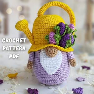 May include: A crocheted gnome wearing a yellow watering can hat with purple flowers. The gnome is sitting on a white surface with white and purple flower petals.
