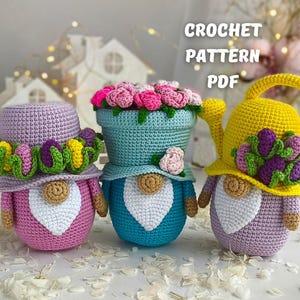 Puede incluir: Tres gnomos de crochet con coronas de flores. El gnomo de la izquierda lleva un sombrero morado con una corona de flores verde y rosa. El gnomo del medio lleva un sombrero azul con una corona de flores rosa. El gnomo de la derecha lleva un sombrero amarillo con una corona de flores morada y una regadera.