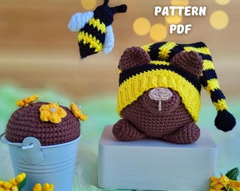 May include: Crochet pattern PDF for a brown bear wearing a yellow and black striped hat. The bear is sitting on a light blue platform. There is a small brown crocheted pot with yellow flowers on top of it. There is also a crocheted bee with black and yellow stripes.