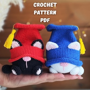 May include: Two crocheted cat plushies wearing graduation caps. One cat is black with a red cap and the other is white with a blue cap. The caps have yellow tassels. The text "CROCHET PATTERN PDF" is visible above the plushies.