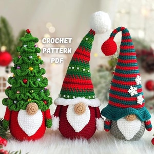 May include: Three crocheted Christmas gnomes, one with a green and red striped hat, one with a red hat, and one with a green Christmas tree hat. The gnomes are sitting on a white surface with a red and green garland in the foreground.