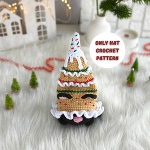 Pode incluir: Um chapéu de árvore de Natal de crochê com um design branco, vermelho e verde. O chapéu tem uma base marrom com uma borda branca semelhante a glacê e um rosto preto com uma boca rosa. O texto "ONLY HAT CROCHET PATTERN" está em um círculo branco.