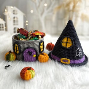 Op de afbeelding: Een Halloween-display met een gehaakte huisvormige mand gevuld met snoep, een zwarte heksenhoed en kleine gehaakte pompoenen. De hoed heeft een paarse band en een wit spinnenwebontwerp. De mand is grijs.