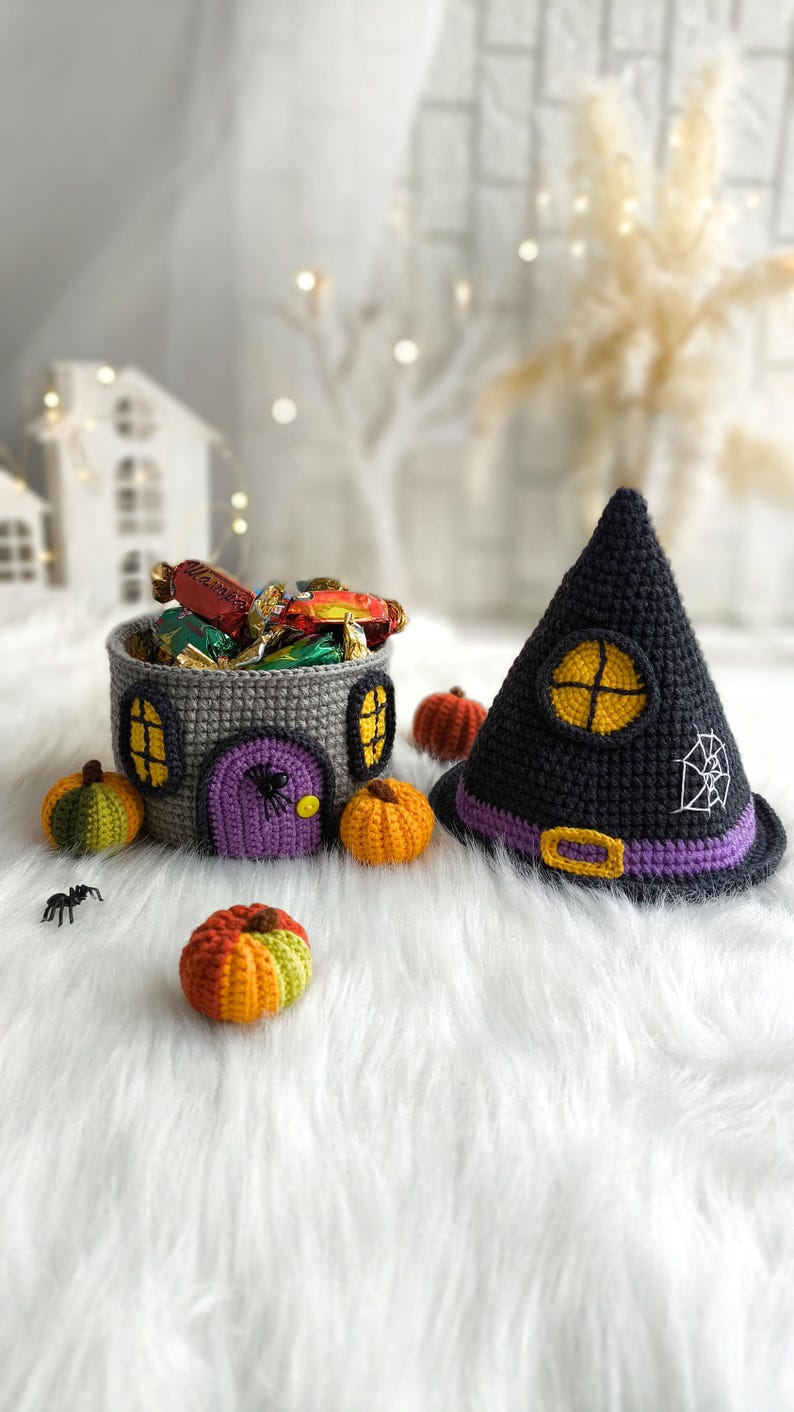 Op de afbeelding: Een Halloween-display met een gehaakte huisvormige mand gevuld met snoep, een zwarte heksenhoed en kleine gehaakte pompoenen. De hoed heeft een paarse band en een wit spinnenwebontwerp. De mand is grijs.
