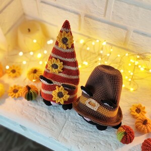 May include: Two crochet gnome figures with sunflower accents. One gnome is tall and conical, with red and white stripes and yellow sunflowers. The other is a brown hat with a yellow band. Small crochet pumpkins and sunflowers are scattered around.