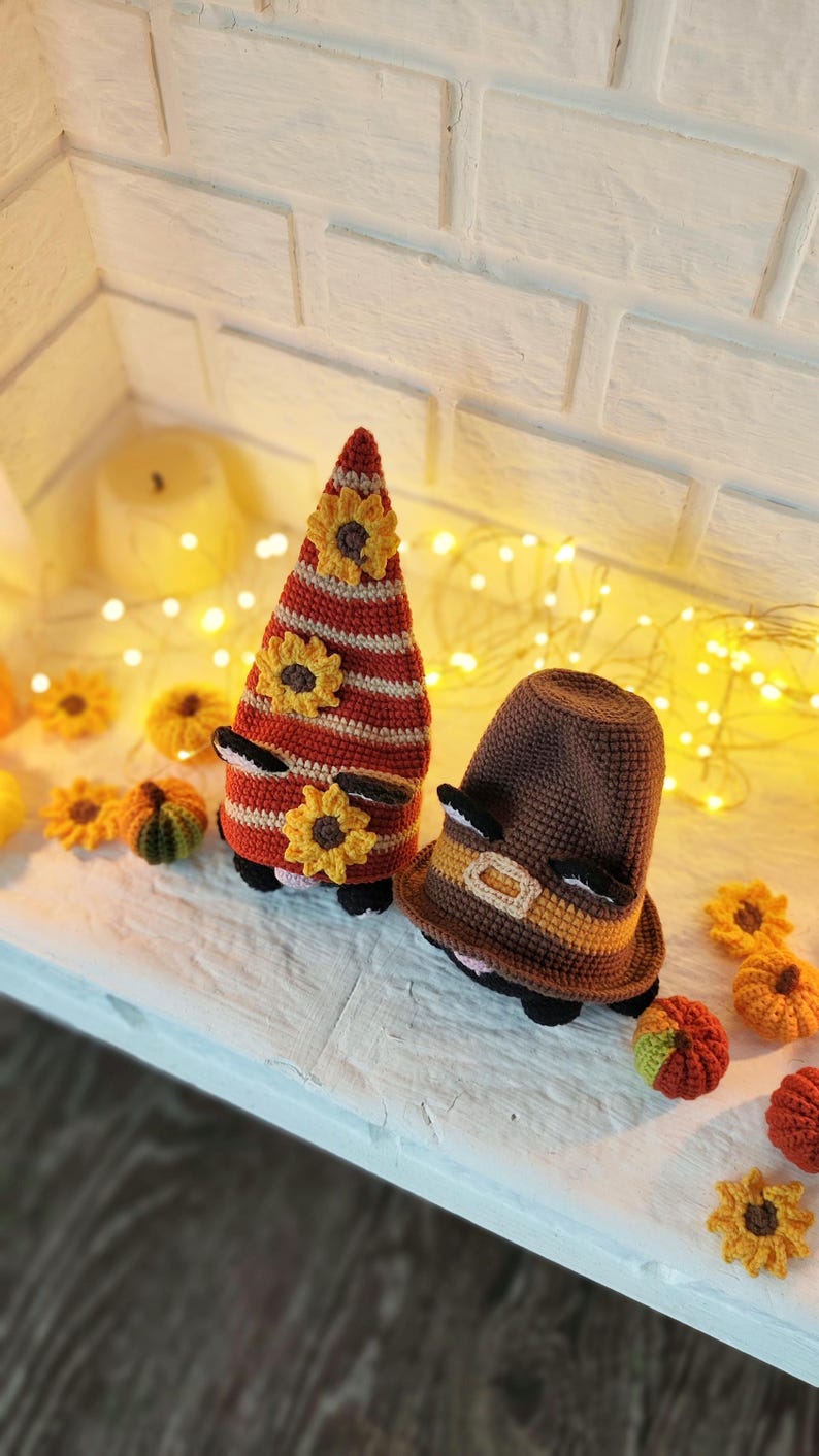 May include: Two crochet gnome figures with sunflower accents. One gnome is tall and conical, with red and white stripes and yellow sunflowers. The other is a brown hat with a yellow band. Small crochet pumpkins and sunflowers are scattered around.