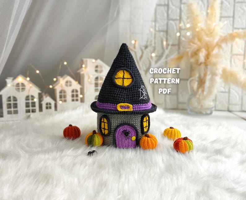 Op de afbeelding: Een gehaakt Halloween-huis met een zwart puntdak, paarse accenten en een geel raam. Rondom het huis liggen kleine gehaakte pompoenen in verschillende kleuren. De tekst "CROCHET PATTERN PDF" is zichtbaar.