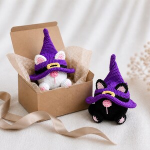 May include: Two crochet cat figurines wearing purple witch hats. One white cat is in a brown box with tissue paper, and the other is a black cat. The hats have black bands and yellow buckles. The cats have pink noses and ears.