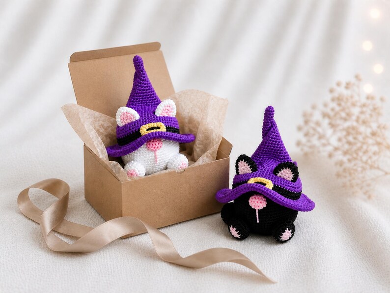 May include: Two crochet cat figurines wearing purple witch hats. One white cat is in a brown box with tissue paper, and the other is a black cat. The hats have black bands and yellow buckles. The cats have pink noses and ears.