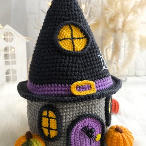 Op de afbeelding: Een gehaakt Halloween-huis met een zwart heksenhoeddak, grijze muren en een paarse deur. Het huis heeft gele raamkozijnen en is versierd met een spin, pompoenen en een paarse riem. Het huis is ongeveer 15 cm hoog.