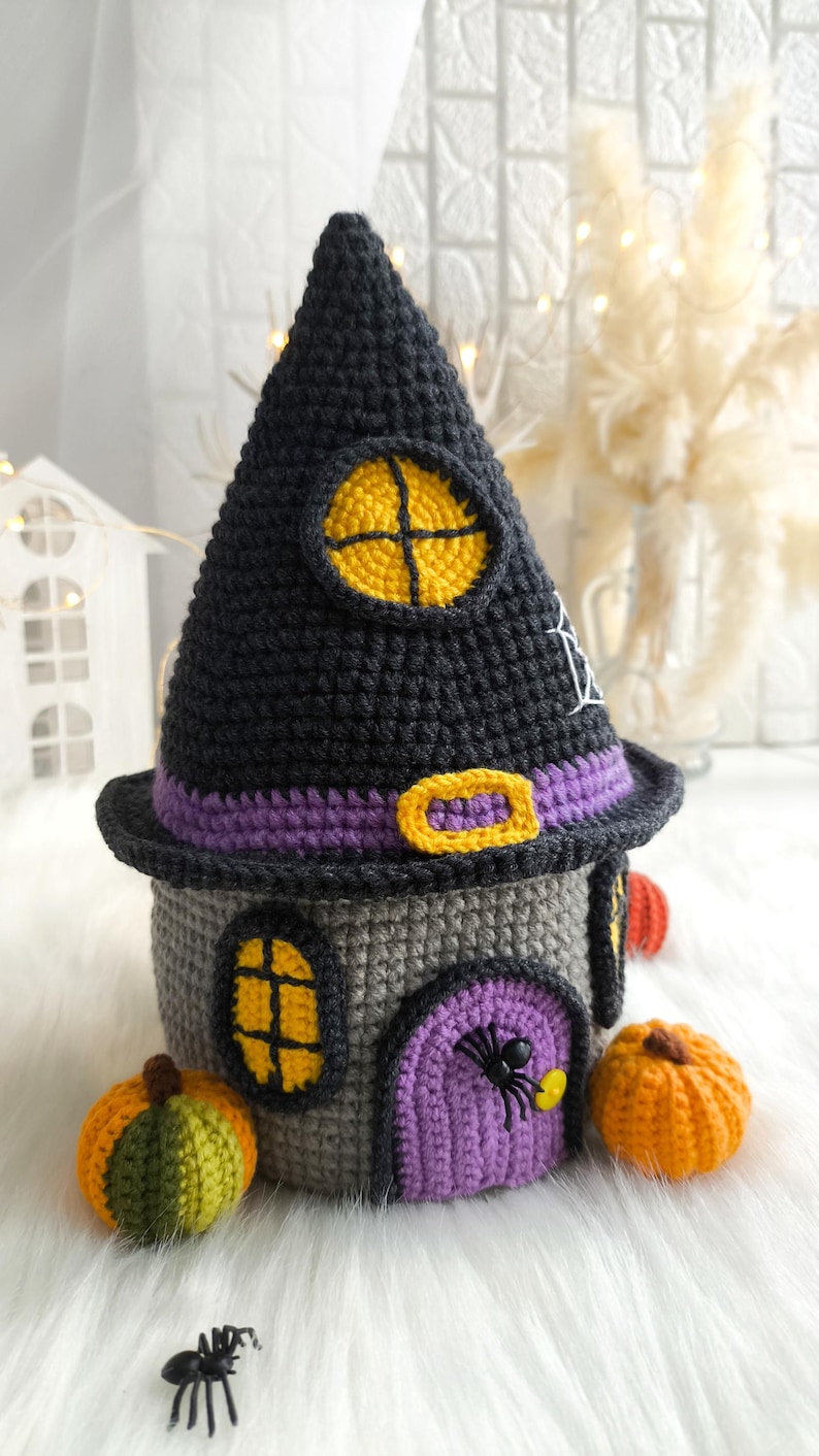 Op de afbeelding: Een gehaakt Halloween-huis met een zwart heksenhoeddak, grijze muren en een paarse deur. Het huis heeft gele raamkozijnen en is versierd met een spin, pompoenen en een paarse riem. Het huis is ongeveer 15 cm hoog.