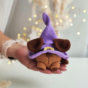 May include: A small crocheted brown dog wearing a purple witch hat with a yellow brim. The dog is sitting in the palm of a hand.
